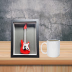 Red electric guitar in a black frame on a wooden surface with a white mug.